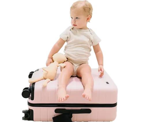 A smiling baby, holding a stuffed bear, seated on top of a pink suitcase