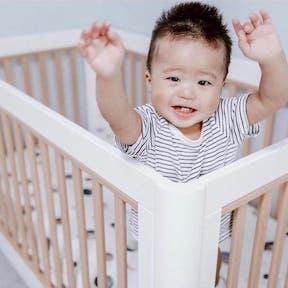 Asian baby boy, standing in crib, arms reaching towards the viewer